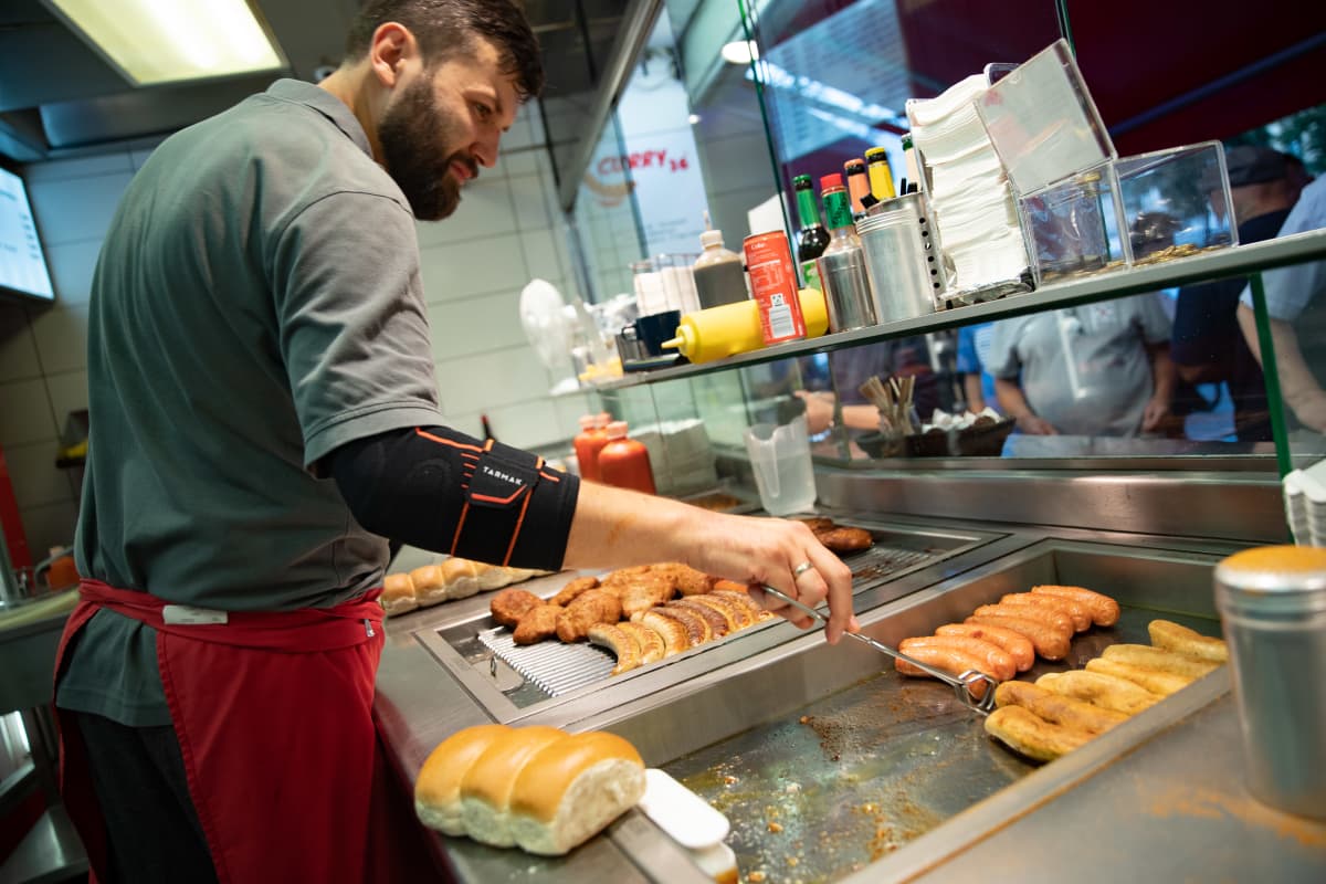 Pikaruokakioskin työntekijä paistaa nakkeja hot dogeihin, kuva on Berliinistä.