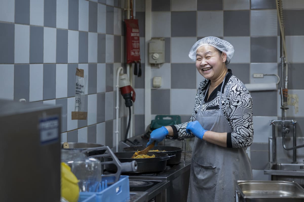 My Duyen Ho sekoittaa hymyillen paistinpannulla paistuvaa lihapiirakoiden täytettä Karjalaisen kotileipomossa. Hänellä on päässään suojamyssy ja kädessä kumihansikkaat. 