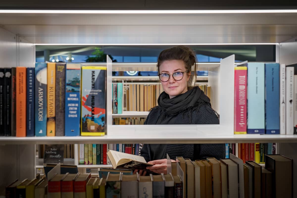 Kääntäjä Riina Behl katsoo kirjahyllyn läpi kohti kameraa.