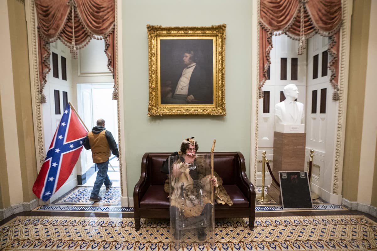 Mielenosoittaja kantaa Etelävaltioiden lippua Yhdysvaltain kongressitalon sisätiloissa. Etualalla istuu turkiksiin pukeutunut henkilö mellakkakilven kanssa.