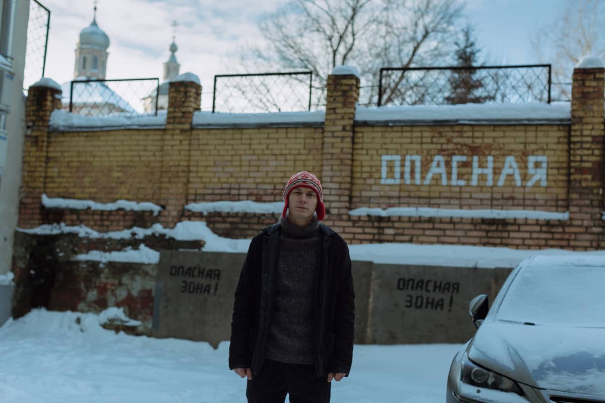 Näyttelijä Juri Borisov seisoo pihalla, taustalla aita ja kirkon tornit, vieressä auto.
