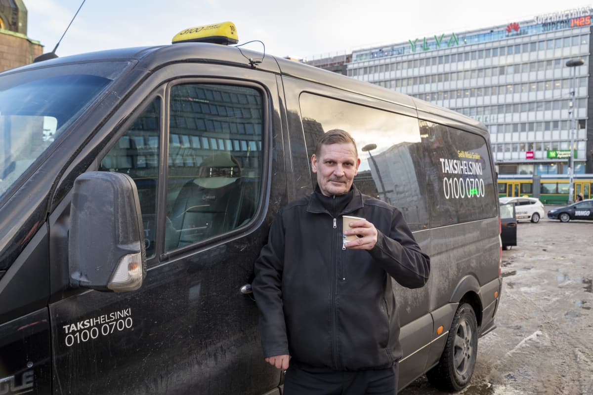 Taksinkuljettaja Lasse Liuski nojailee autoonsa kahvikuppi kädessä.
