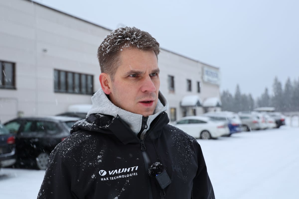 Mies seisoo lumisateessa Vauhti Speed -yhtiön musta takki yllään. Taustalla vaaleaseinäinen tehdas.