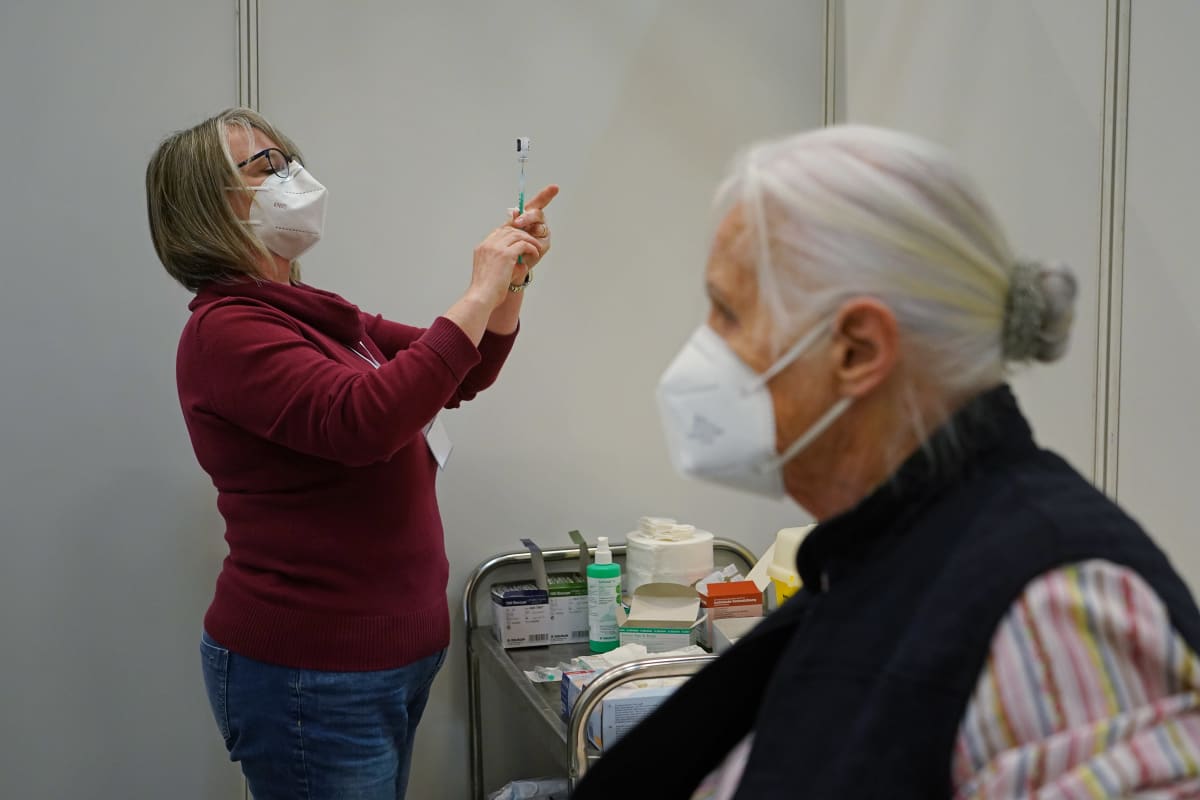 Etualalla iäkäs nainen hengityssuojain kasvolla ja taustalla naispuolinen sairaanhoitaja valmistelee koronarokotetta.