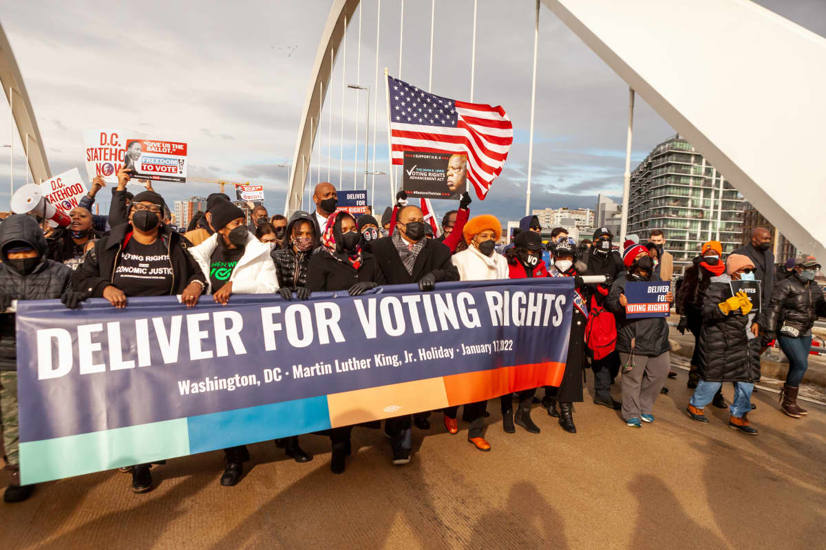 Joukko lähinnä mustia mielenosoittajia kantaa lakanaa, jossa lukee "Deliver for Voting Rights". 