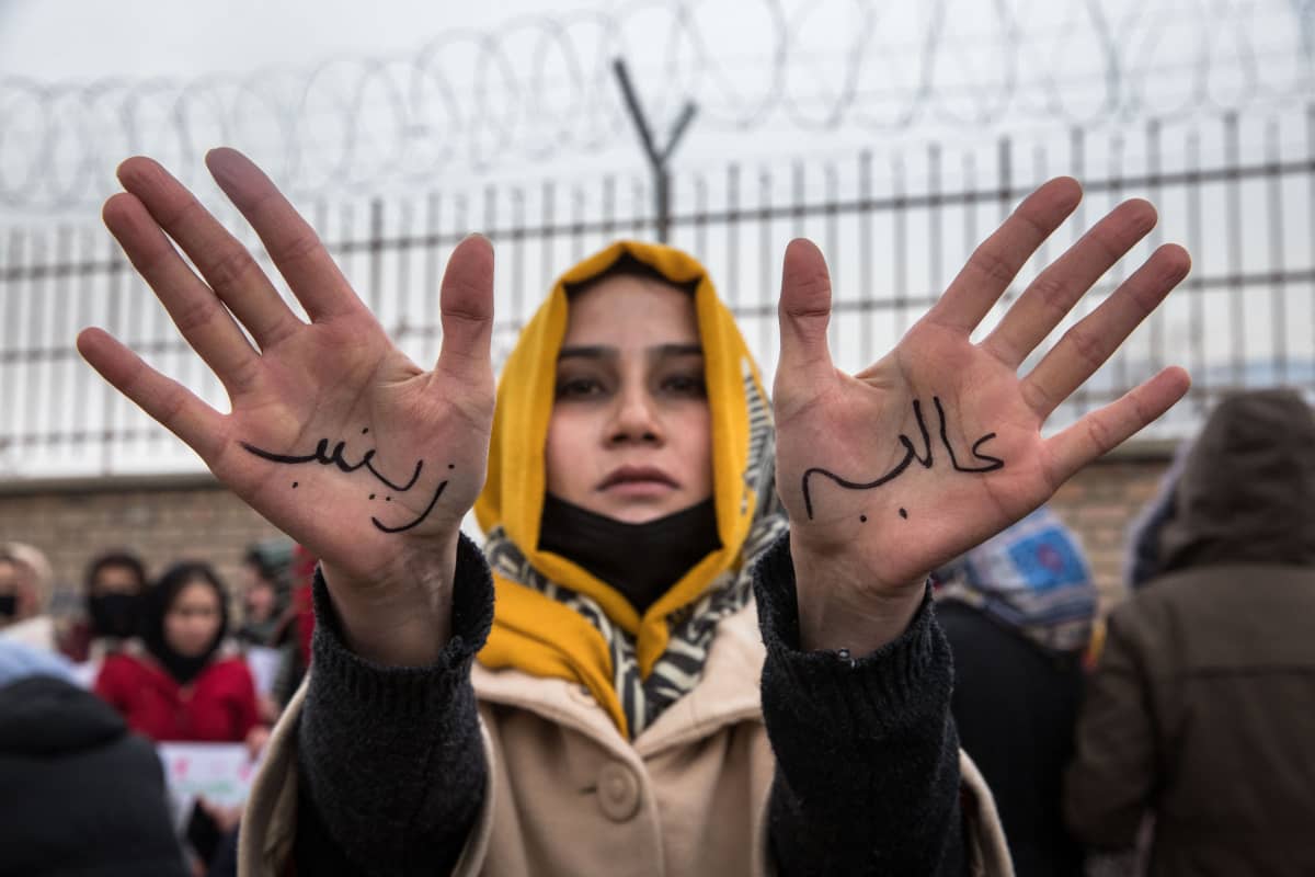 Huiviin pukeutunut nainen näyttää käsiään kameralle mielenosoituksessa. Käsiin on kirjoitettu iskulauseita.