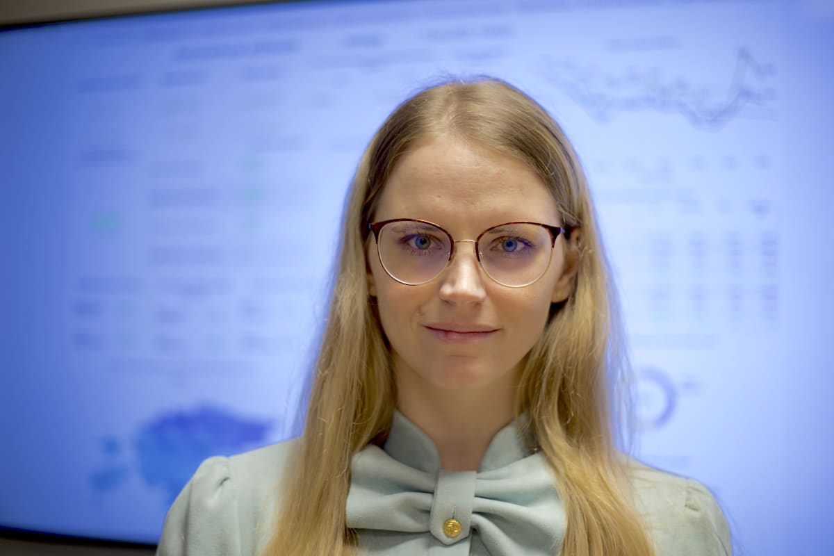Silmälasipäinen nuori vaaleatukkainen nainen hymyilee kameraan takanaan tilastotietoa Viron koronatilanteesta.