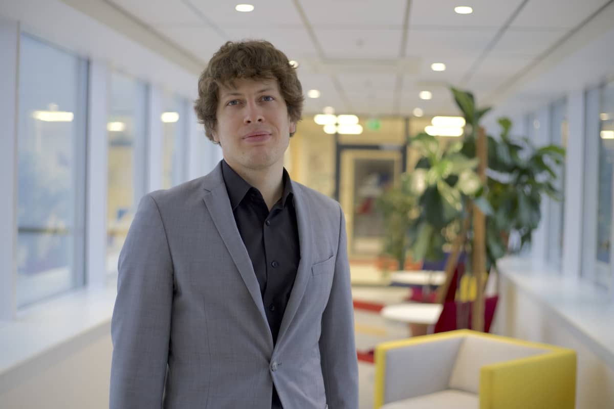 Mies tummassa paidassa ja harmaassa pikkutakissa katsoo suoraan kameraan.