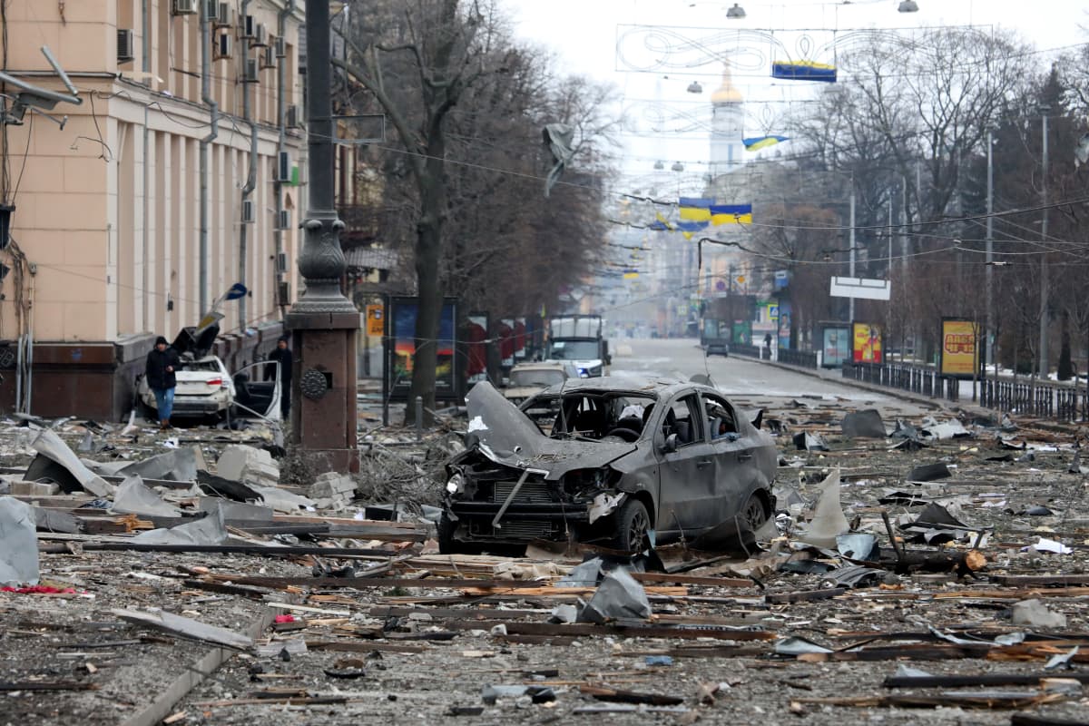 Pommi-iskun jälkiä, tuhoutuneita autoja, taustalla kadulla näkyy Ukrainan lippuja. Ukrai