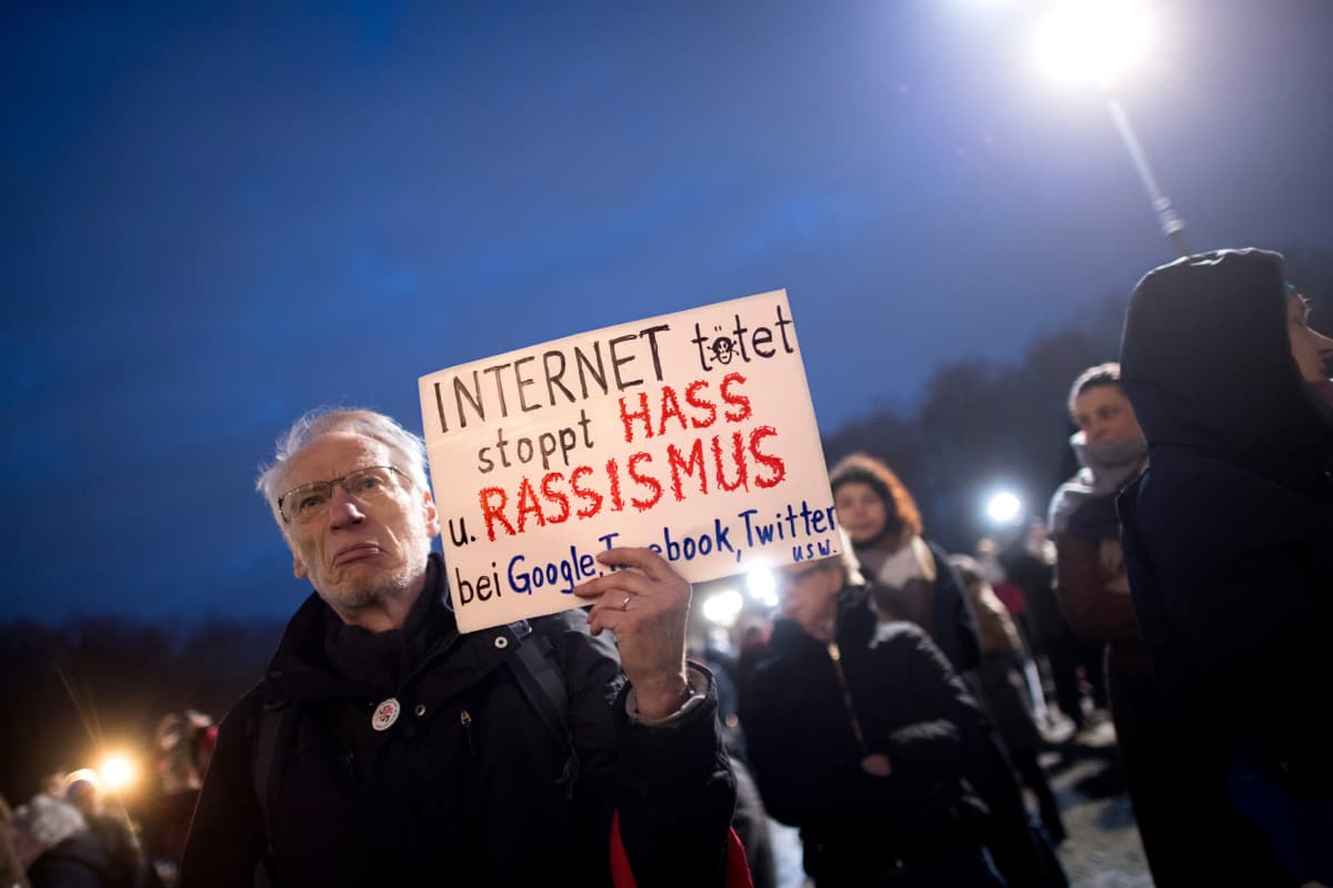 Vihaisen ja samalla surullisen näköinen vanha mies kantaa kylttiä illan pimetessä. Ympärillä on muita mielenosoittajia.