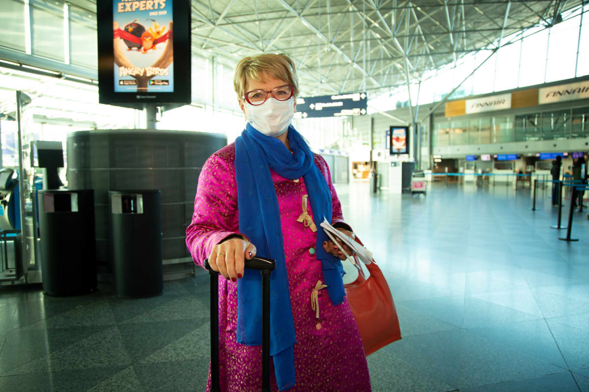 Liisa Hakala Helsinki-Vantaa lentoaseman lähtöaulussa. Liisalla on purppuran punainen takki, jossa pieniä kultaisia kuvioita ja sininen pitkä kaulaliina. Matkatavaroina on vedettävä pieni matkalaukku ja punainen käsilaukku. Kasvoilla on hengityssuojain. 