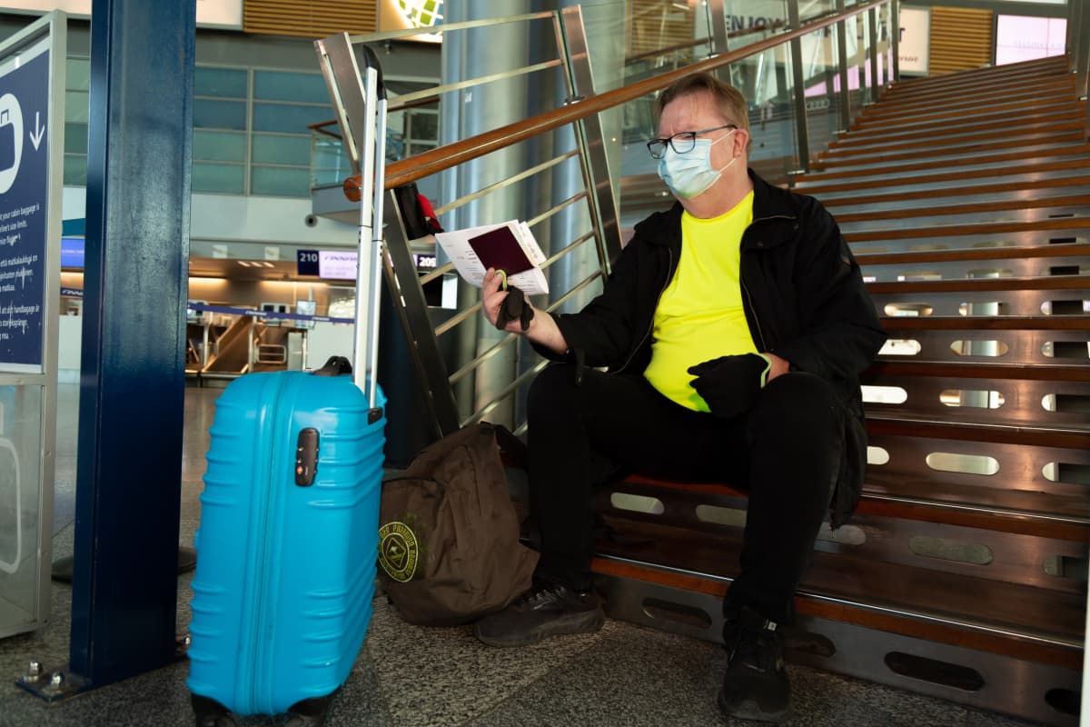 Risto Niemi istuu Helsinki-Vantaan lentoaseman lähtöaulassa rappusilla hengähtämässä ja tarkastamassa matkapapereitaan. Ristolla on keltainen t-paita ja mustat farkkuhousut ja -takki. Vedettävä matkalaukku on kirkkaan sininen.