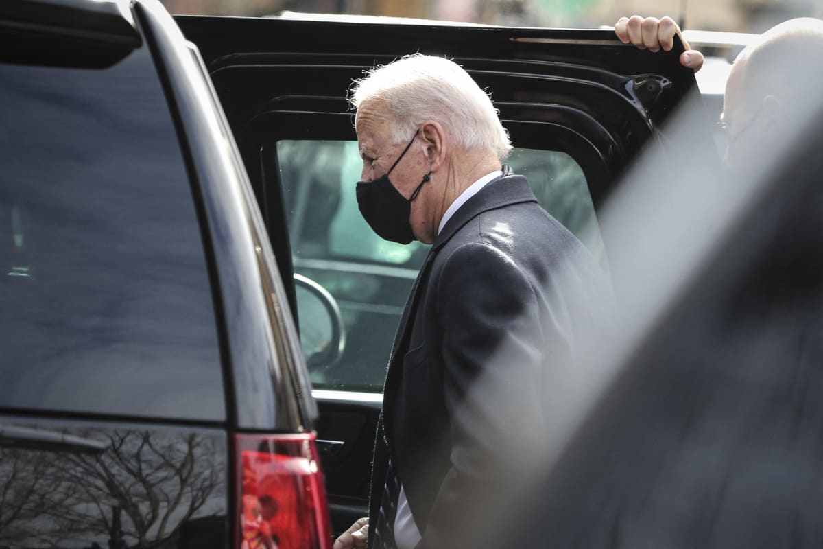 Joe Biden nousee suureen mustaan autoon. Turvamies pitelee ovea auki. Bidenilla on tumma puku ja musta kasvomaski.