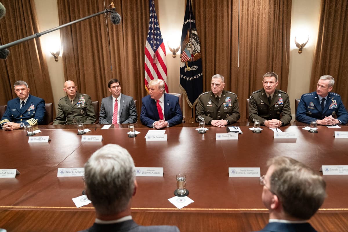 Trump istuu suuren pöydän ääressä, ympärillään amerikkalaisia kenraaleja eri aselajeista erilaisissa univormuissa. 