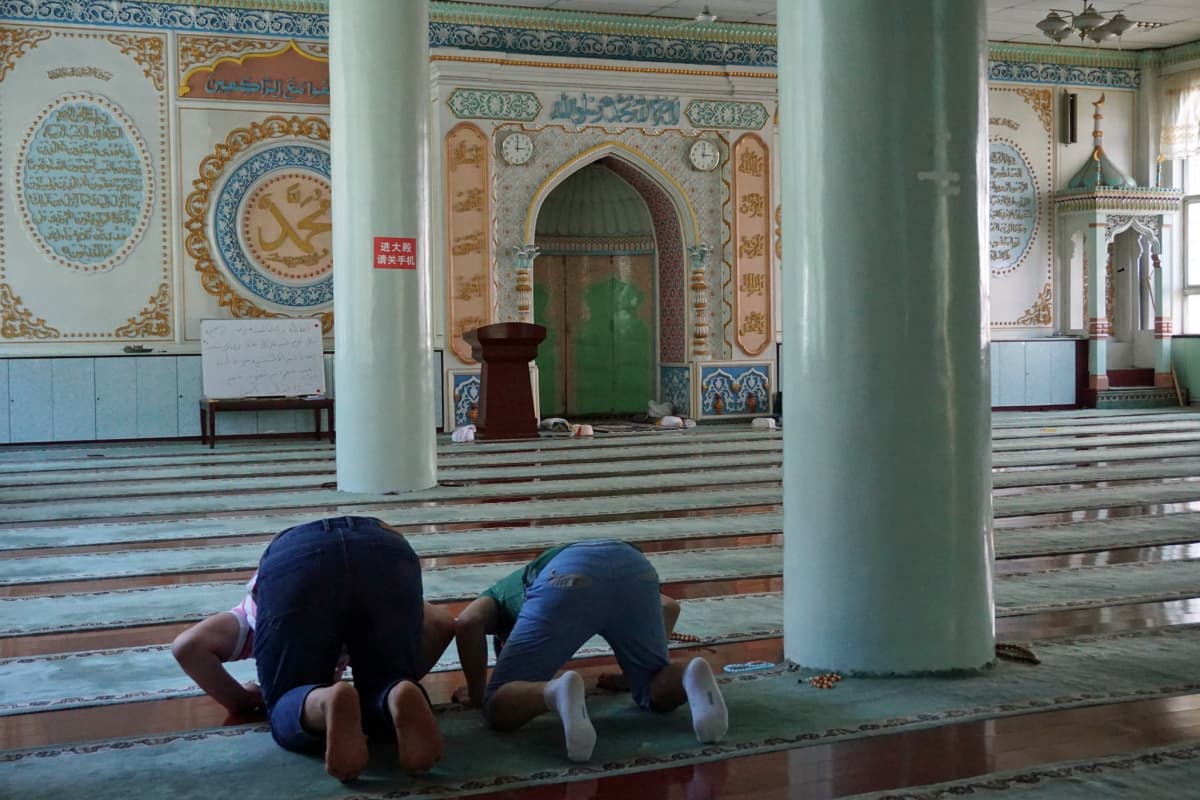 Kuvassa moskeijan vaaleanvihreät pylväät ja koristeellinen seinä, etualalla kaksi rukoilijaa.