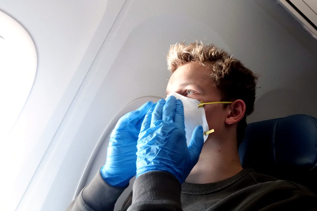 Hengityssuojaimeen ja suojakäsineisiin pukeutunut mies matkustaa lentokoneessa.