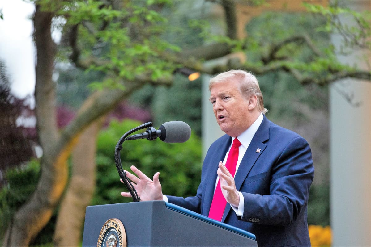 Yhdysvaltain presidentti Donald Trump puhuu mikrofoniin puhujanpöntön takana. Hänellä on tummansininen puku, napinlävessä Yhdysvaltain lippua esittävä pinssi, kauluspaita ja ja kirkkaanpunainen kravatti. Hän korostaa puhettaan molemmilla käsillään.