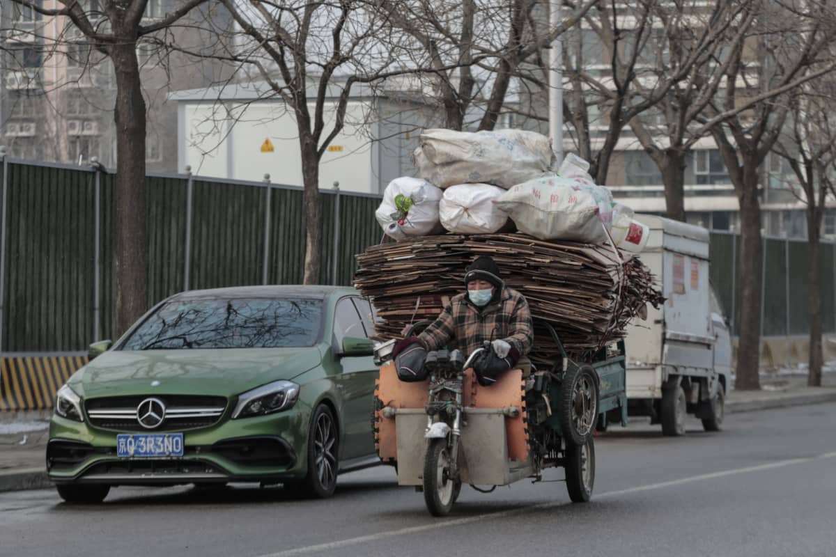 Talvivaatteisiin ja kasvomaskiin pukeutunut mies kuljettaa huojuvaa pahvi- ja kierrätysjätepinoa mopokärryllä.