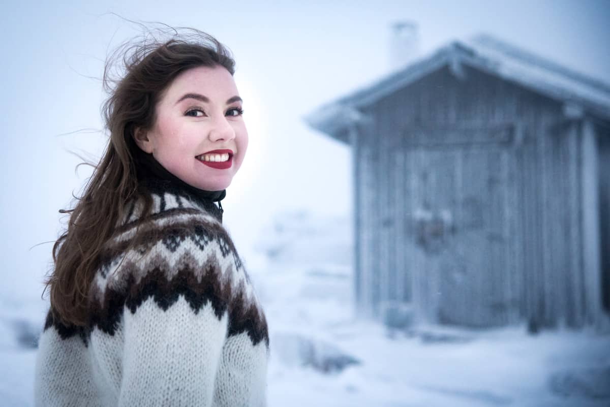 Riikka Karppinen hymyilee punatuilla huulillaan kameralle lumisessa maisemassa ladon edessä päällään valko-ruskea islanninvillapaita.