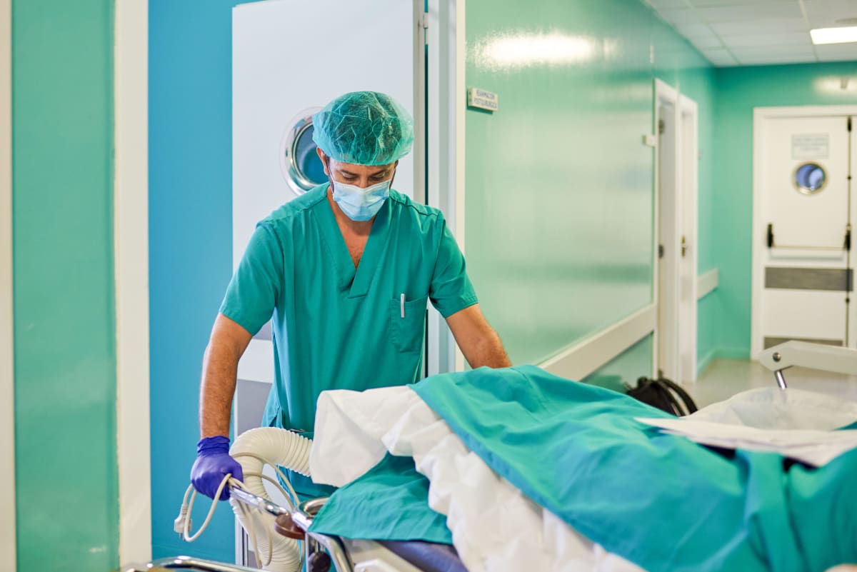 En sjukskötare i munskydd rullar runt en sjukhussäng i en korridor.