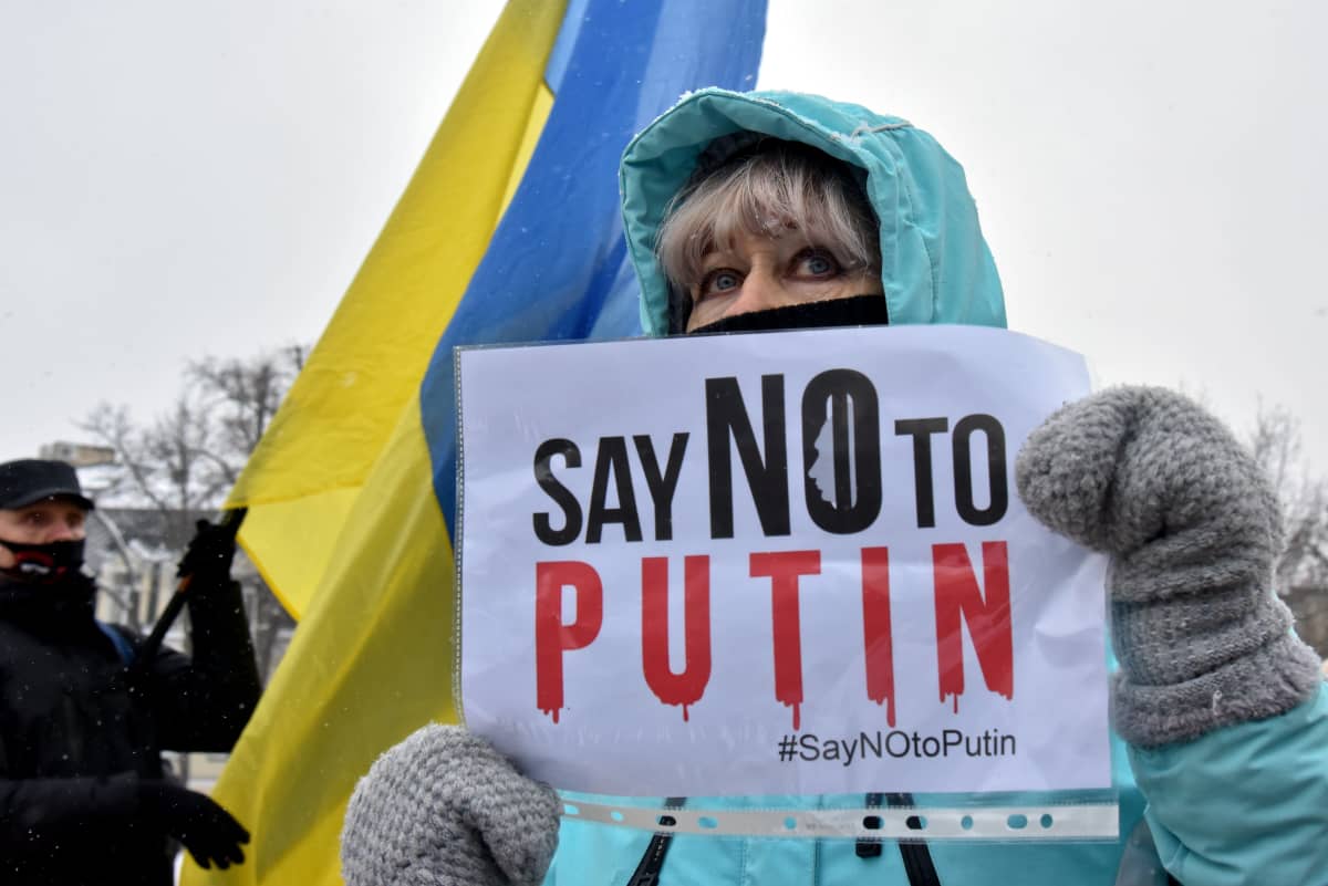 Nainen pitää käsissään kylttiä, jossa lukee "Say no to Putin". Taustalla näkyy keltasininen Ukrainan lippu.
