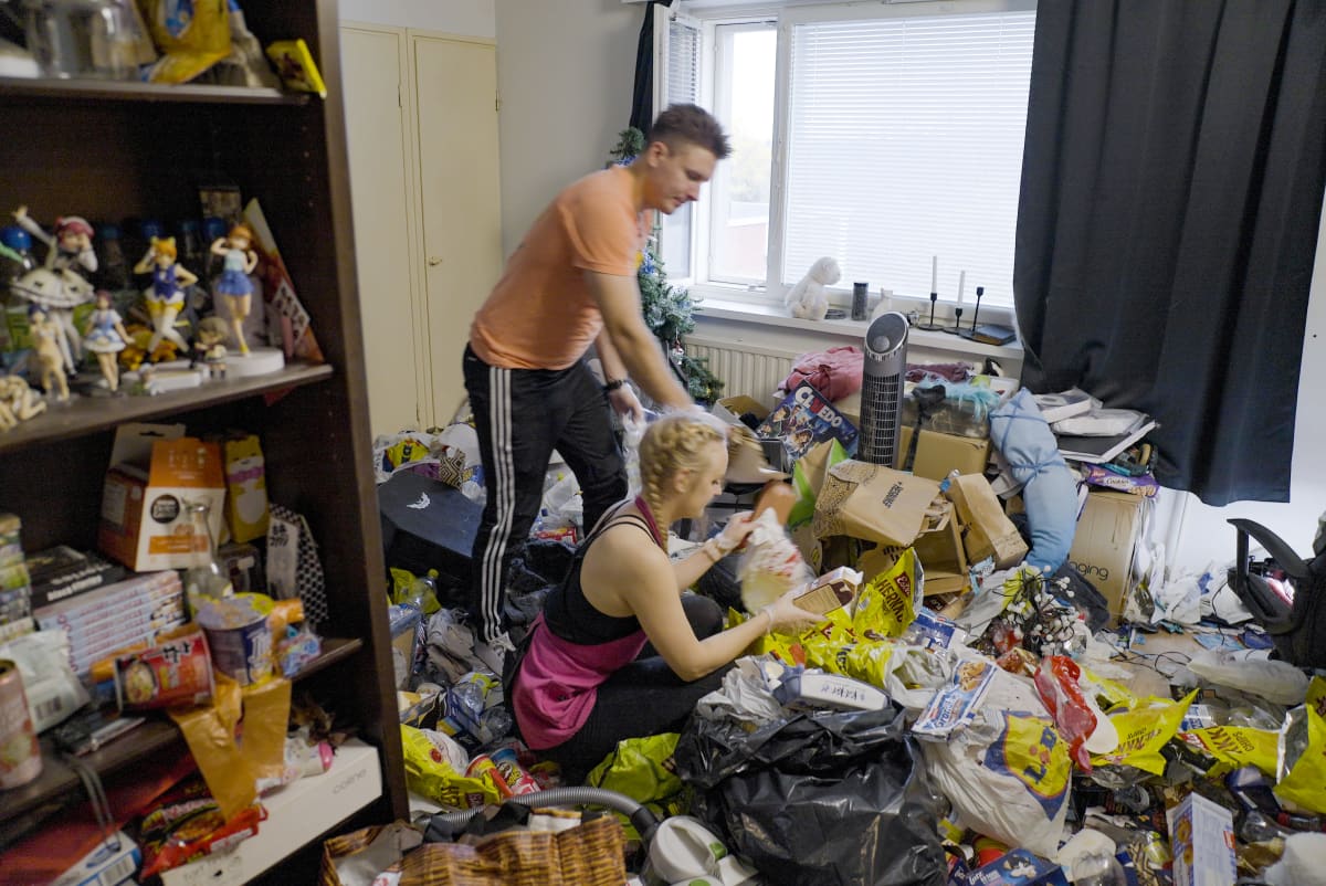 Nuori mies ja nuori nainen keräävät roskia muovipusseihin asunnossa, jossa on niin paljon roskaa, että se peittää lattian.