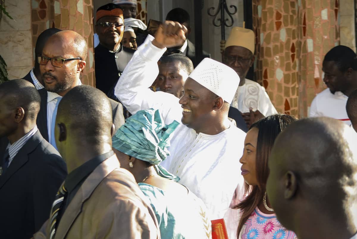 Valkoisiin pukeutunut hymyilevä Adama Barrow heiluttaa kättään. Ympärillä on useita ihmisiä.