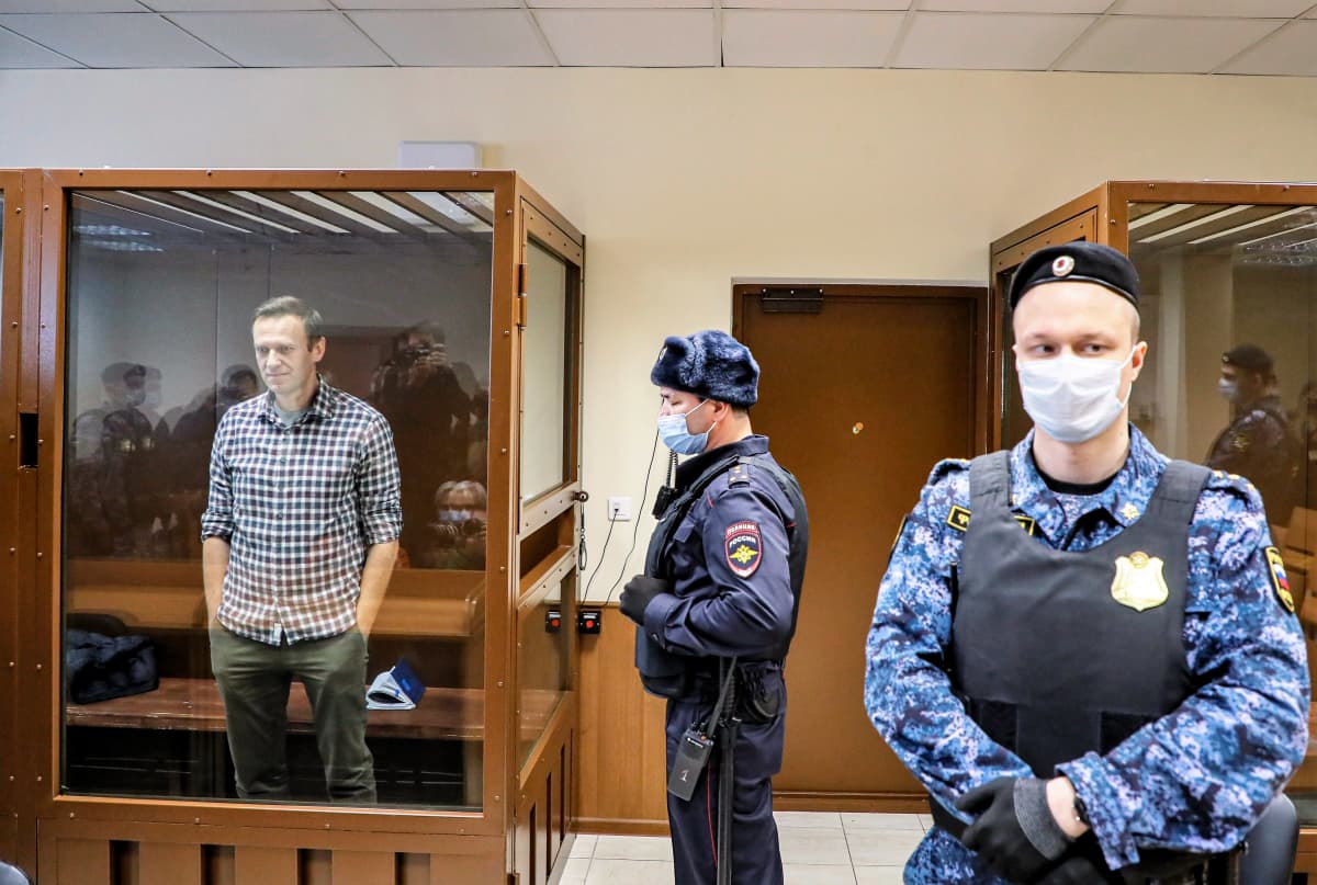 Navalnyi seisoo lasiseinäisessä häkissä oikeussalissa, ruutupaidassa, kädet taskuissa. Häkin ulkopuolella seisoo kaksi univormupukuista miestä vartiossa.