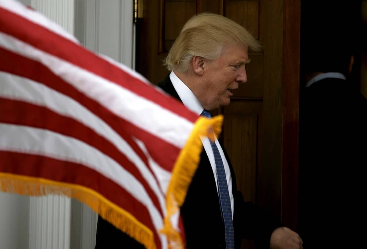 Donald Trum kulkemassa ovesta. Edessä liehuu Yhdysvaltojen lippu.