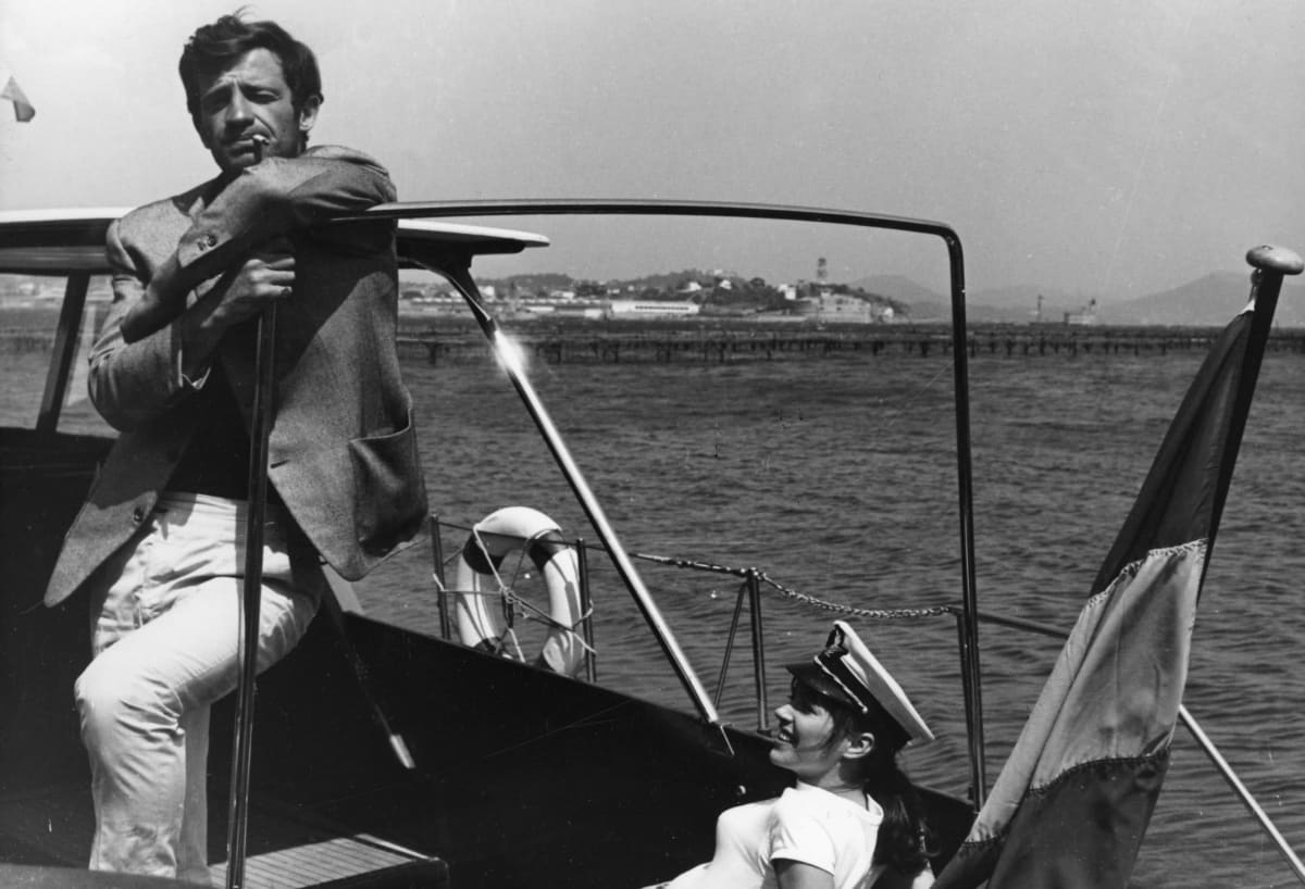 Belmondo seisoo veneessä tupakka suupielessään ja katsoo kohti kameraa. Veneessä istuu Anna Karina kapteeninlakki päässään.