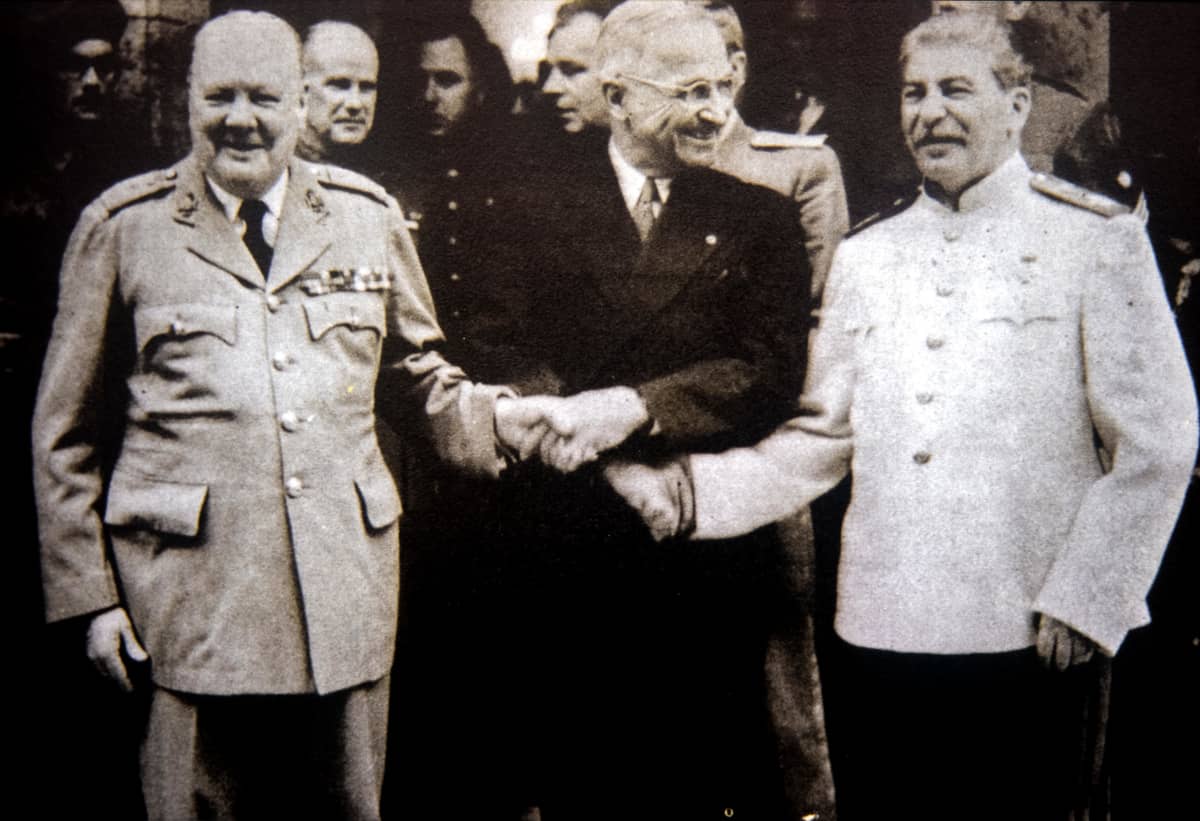 Winston Churchill, Harry S. Truman ja Josef Stalin pitävät toistensa käsistä kiinni. Churchillillä ja Stalinilla on univormut, Trumanilla tumma puku.