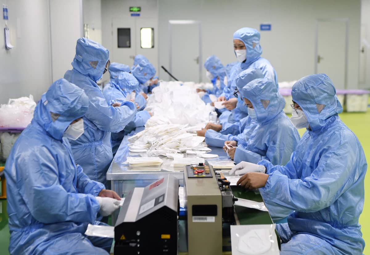 Sinisiin suojavarusteisiin ja valkoisiin kasvosuojaimiin pukeutuneet työläiset valmistelevat kasvosuojaimia pöydän ääressä.