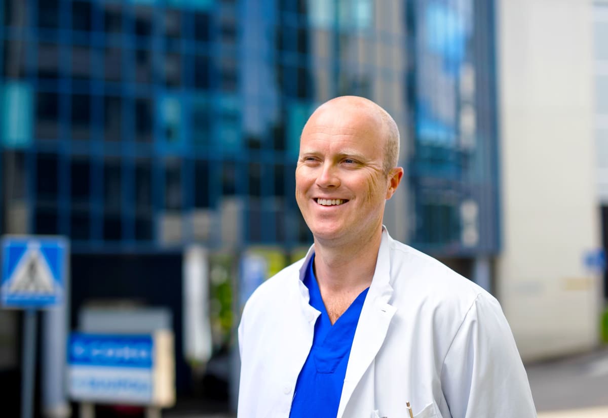 Ville Mattila seisoo sairaalan ulkopuolella valkoinen lääkärintakki yllään.
