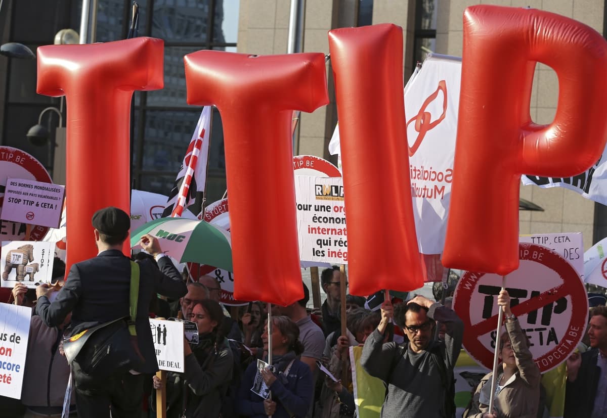 Kuvassa isot, punaiset ilmapallokirjaimet jotka muodostavat lyhenteen TTIP ja TTIP:tä vastustavia mielenosoittajia.