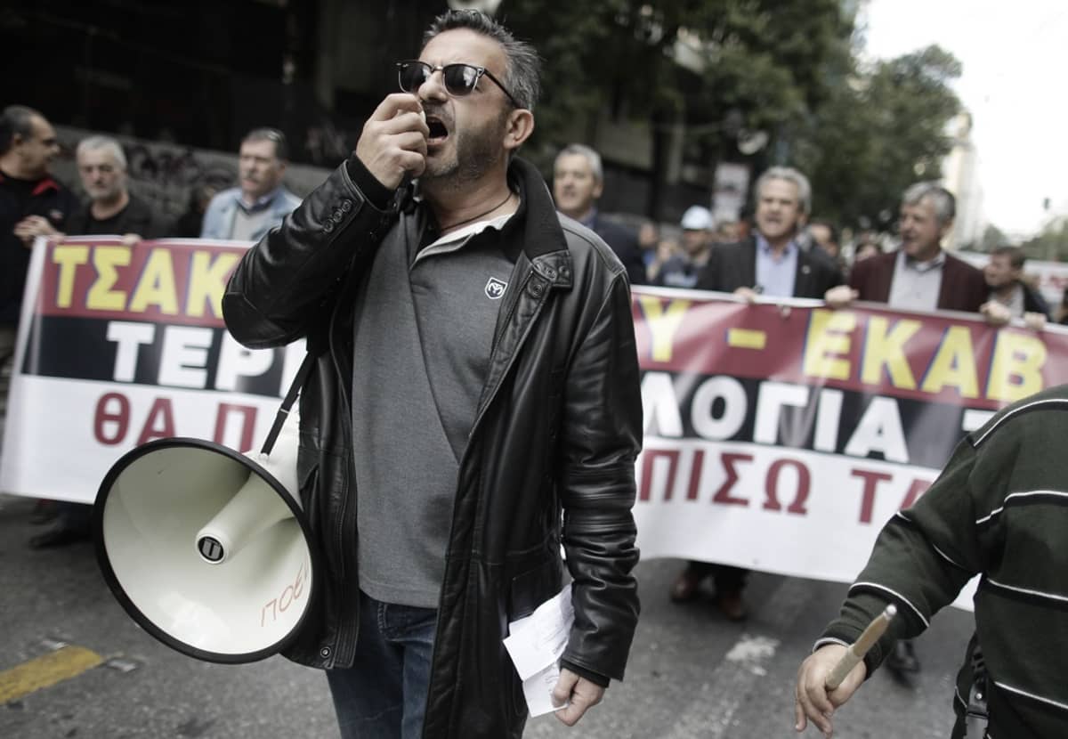 Mielenosoitusta. Etualalla mustaan nahkatakkiin pukeutunut parrakas mies mustissa silmälaseissa puhuu megafonin mikrofoniin. Taustalla joukko mielenosoittajia kantaa plakaattia, jossa on kreikankielistä kirjoitusta.