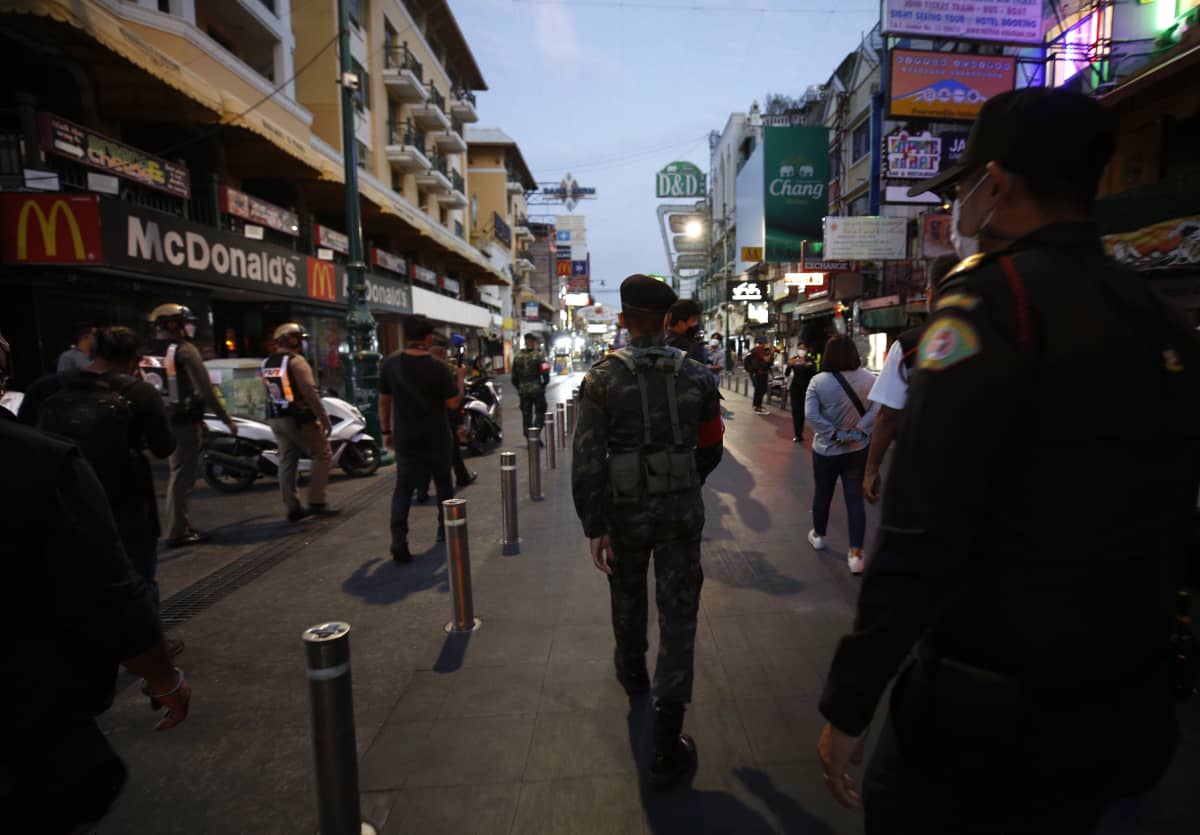 Thaimaalaiset poliisit ja sotilaat kävelevät ostos- ja bilekadulla iltahämärässä. Kadun reunoilla näkyy ravintoloiden kylttejä ja mainoksia.