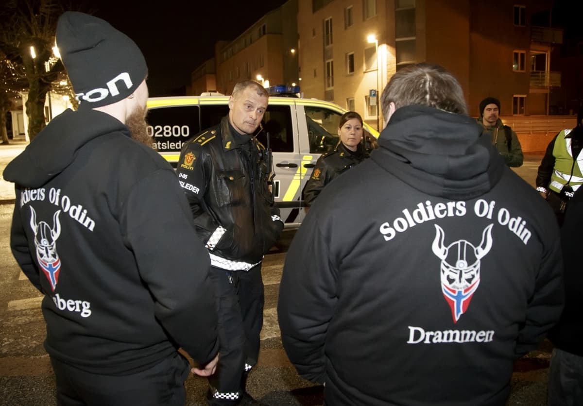 Selin kaksi miestä, joiden mustien pusakoiden selkämuksessä on lukee Soldiers of Odin ja Drammen. Välissä sarvikypärään pukeutunut hahmo, jolla partana Norjan lippu. Takana kaksi poliisia ja poliisiauto. 