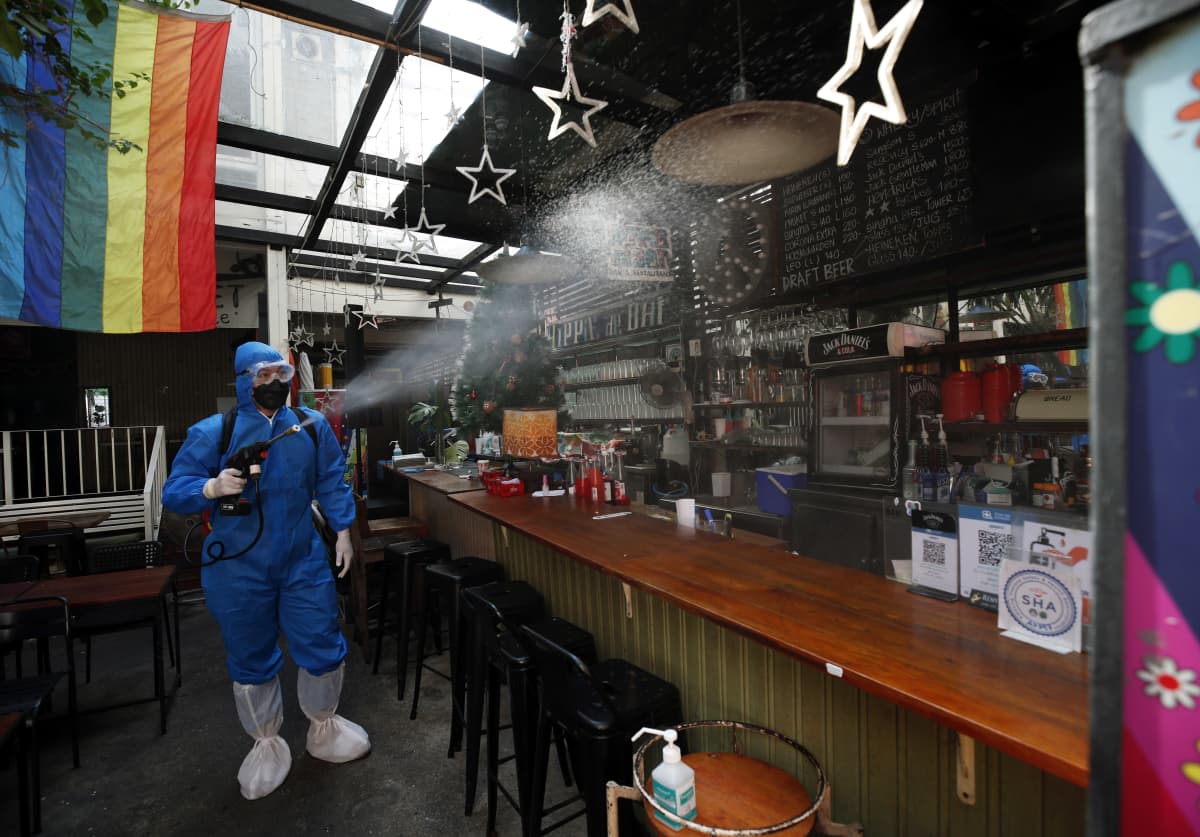 Siniseen suoja-asuun pukeutunut työntekijä suihkuttaa puhdistusainetta baaritiskin ylle tyhjässä yökerhossa. Tiskillä on muovinen joulukuusi ja katosta roikkuu sateenkaarilippu.