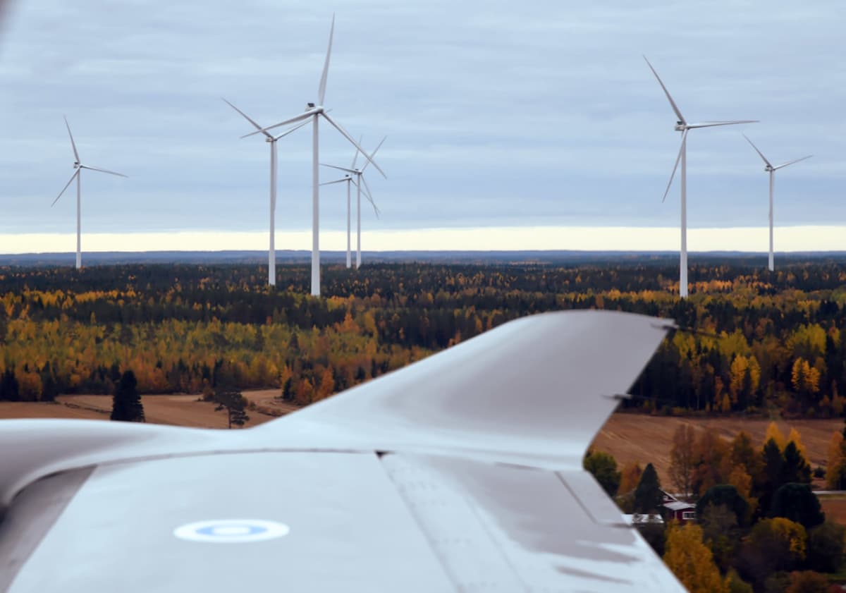 Ilmavoimien Pilatus PC-12 NG -yhteyskoneen siipi, jonka taustalla näkyy tuulivoimaloita syksyisessä maaseudun metsämaisemassa.