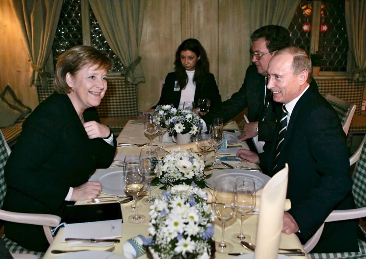 Angela Merkel ja Vladimir Putin hyväntuulisina ruokapöydän ääressä, pöydällä kukka-asetelma, ruokaa ei vielä lautasella. 