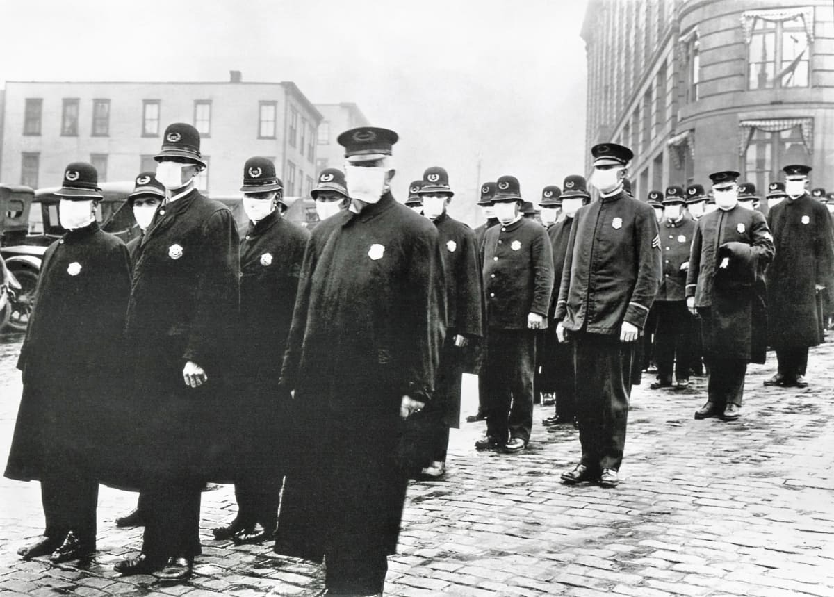 Joukko univormupukuisia poliisimiehiä seisoo kadulla suojaimet kasvoillaan. Kuva on mustavalkoinen.