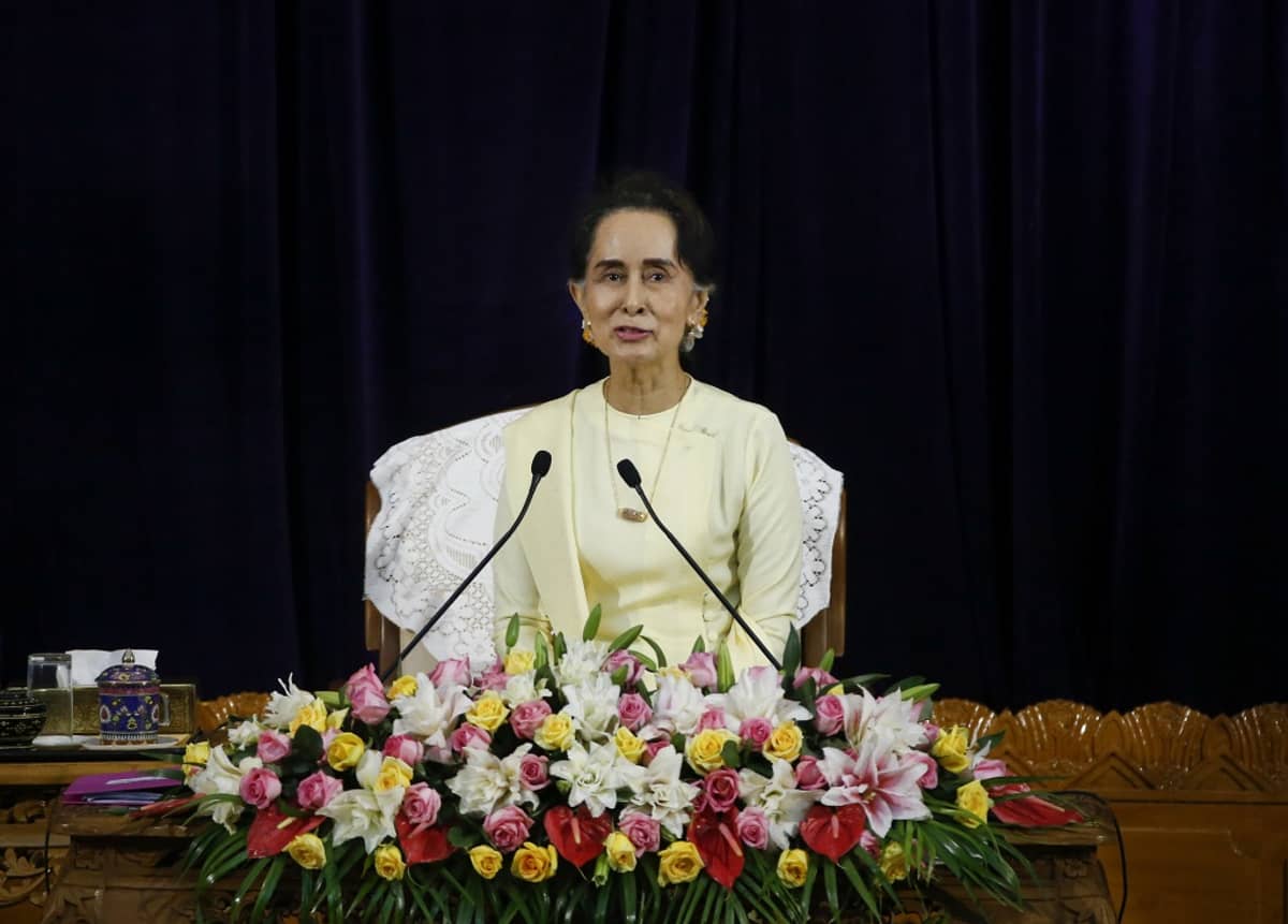 Aung San Suu Kyi istuu kukkalaitteen ja kahden mikrofonin takana vaaleankeltaisessa asussa tummaa taustaa vasten.