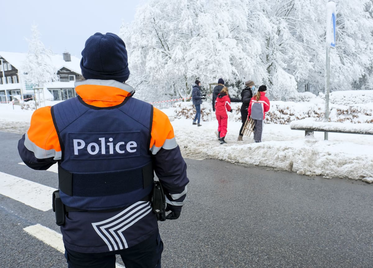 Poliisi seisoo selin kameraan, taustalla joukko ihmisiä mäenlaskuvarusteiden kanssa lumisessa maisemassa
