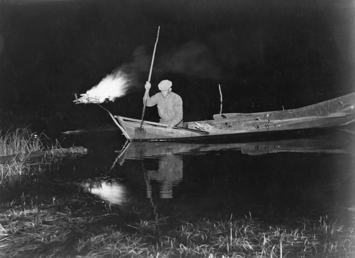 Vanha kuva, jossa mies tuulastaa järvellä. Veneen kokassa palaa iso soihtu. 