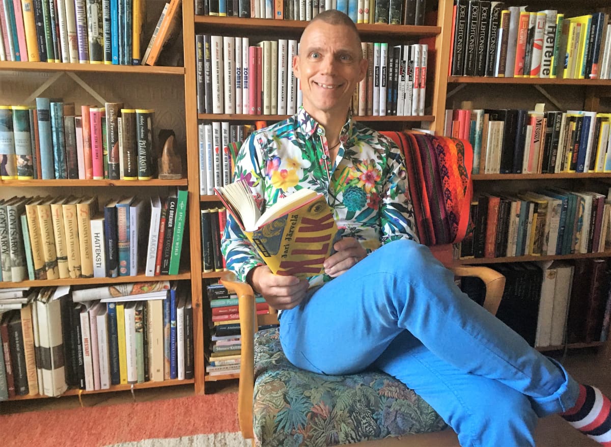 Jari Järvelä istuu tuolissa kirjahyllyn edessä kirja kädessä 