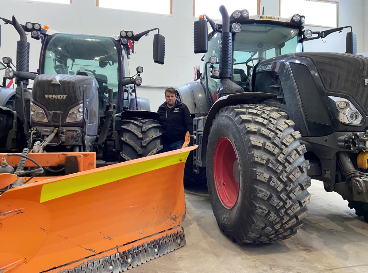 Maatalousyrittäjä Juha-Pekka Toukola seisoo konehallissa kahden ison traktorinsa välissä. Toisen traktorin keulaan on asennettu lumiaura.