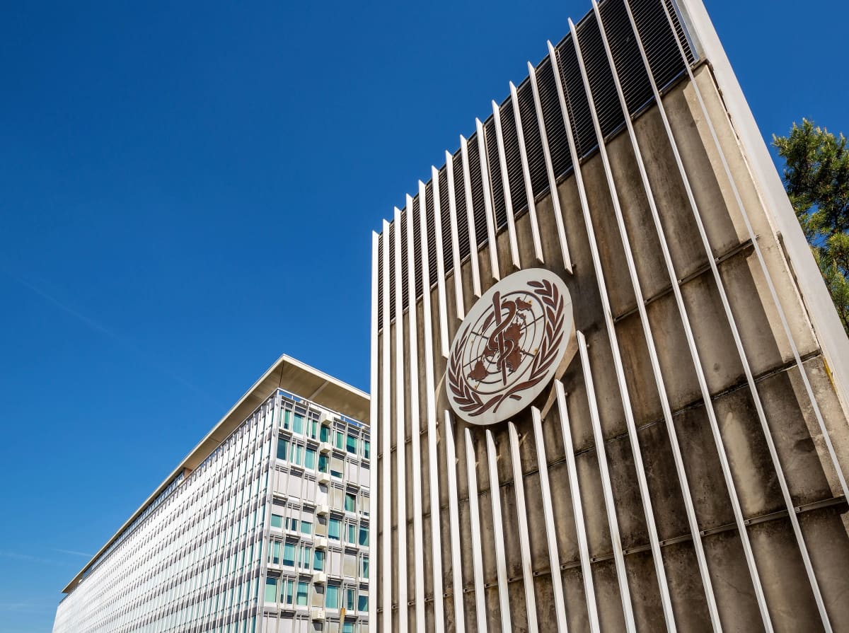 Kuvassa on Maailman terveysjärjestön WHO:n päämaja. Portissa on järjestön logo, jossa on YK:ta kuvaava maapallo ja lehvät ja maapallon keskellä terveydenhuoltoa symboloiva käärme sauvan ympärille kietoutuneena.