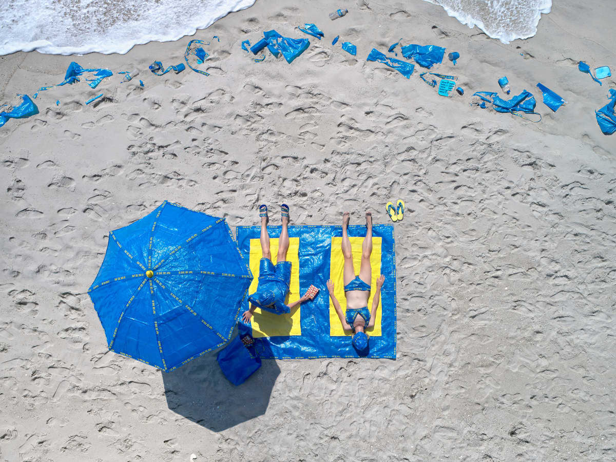 Valokuvateos, jossa Ikea-kasseja auringonvarjoina ja pyyhkeenä käyttävät kaksi ihmistä makaavat rannalla. Rannalla näkyy Ikea-kasseja hiekan seassa.