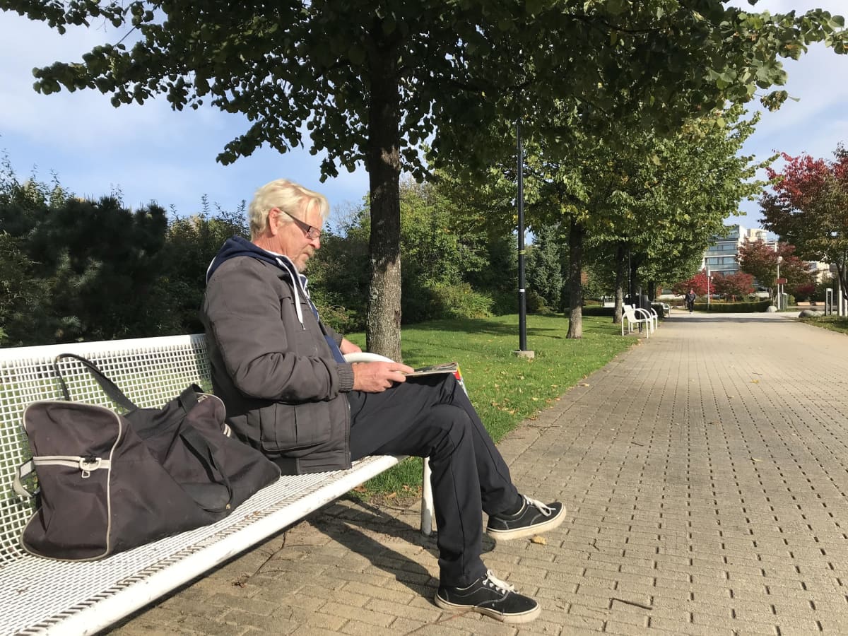 Timo Muhonen lukee lehteä puistossa, hän istuu puun alla penkillä lähellä Keravan kirjastoa.