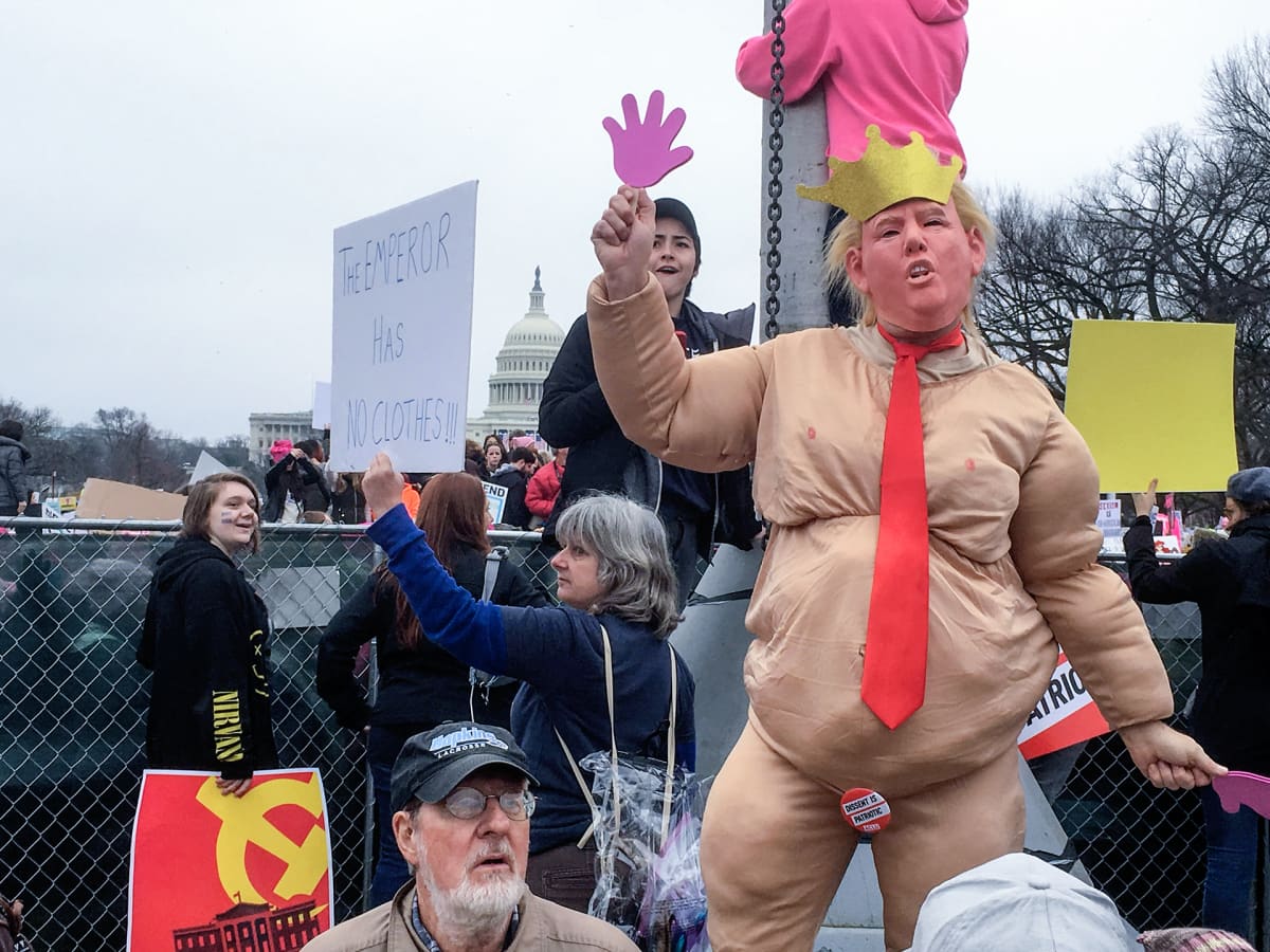 Henkilö on pukeutunut Donald Trump-naamariin ja pukuun, joka saa hänet näyttämään lihavalta ja alastomalta. Nainen pitää kylttiä, jossa lukee "The emperor has no clothes."